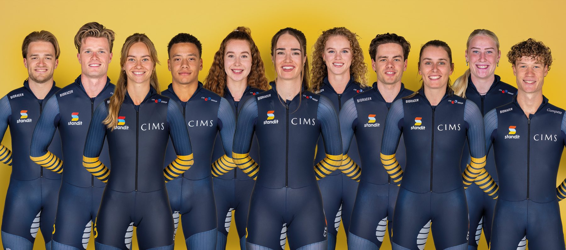 De elf schaatsers van Team Staan CTS GROUP naast elkaar in teampak, tegen een gele achtergrond